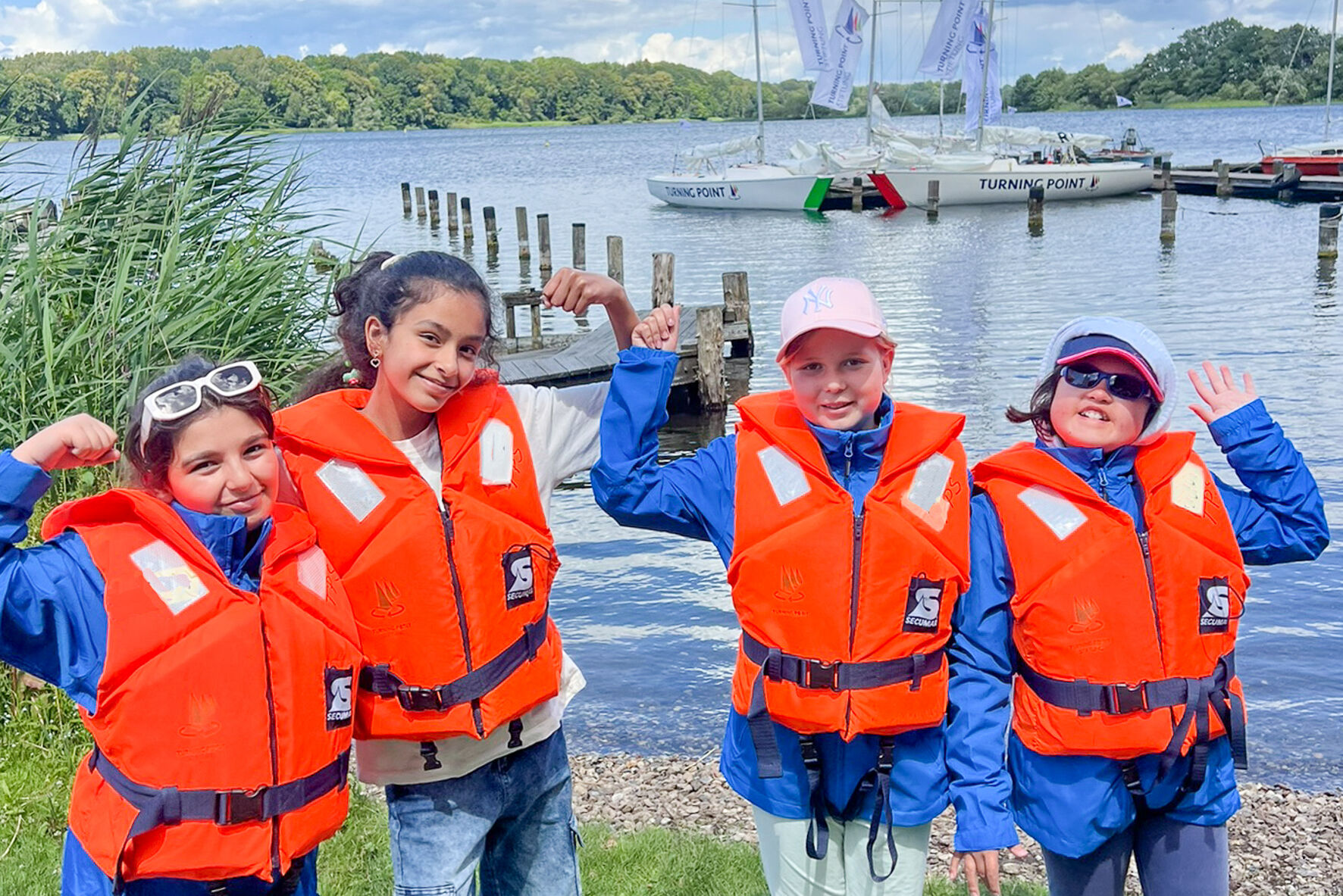 Vier Kinder in Schwimmwesten stehen vor einem See. Sie winken und zeigen ihre starken Arme.