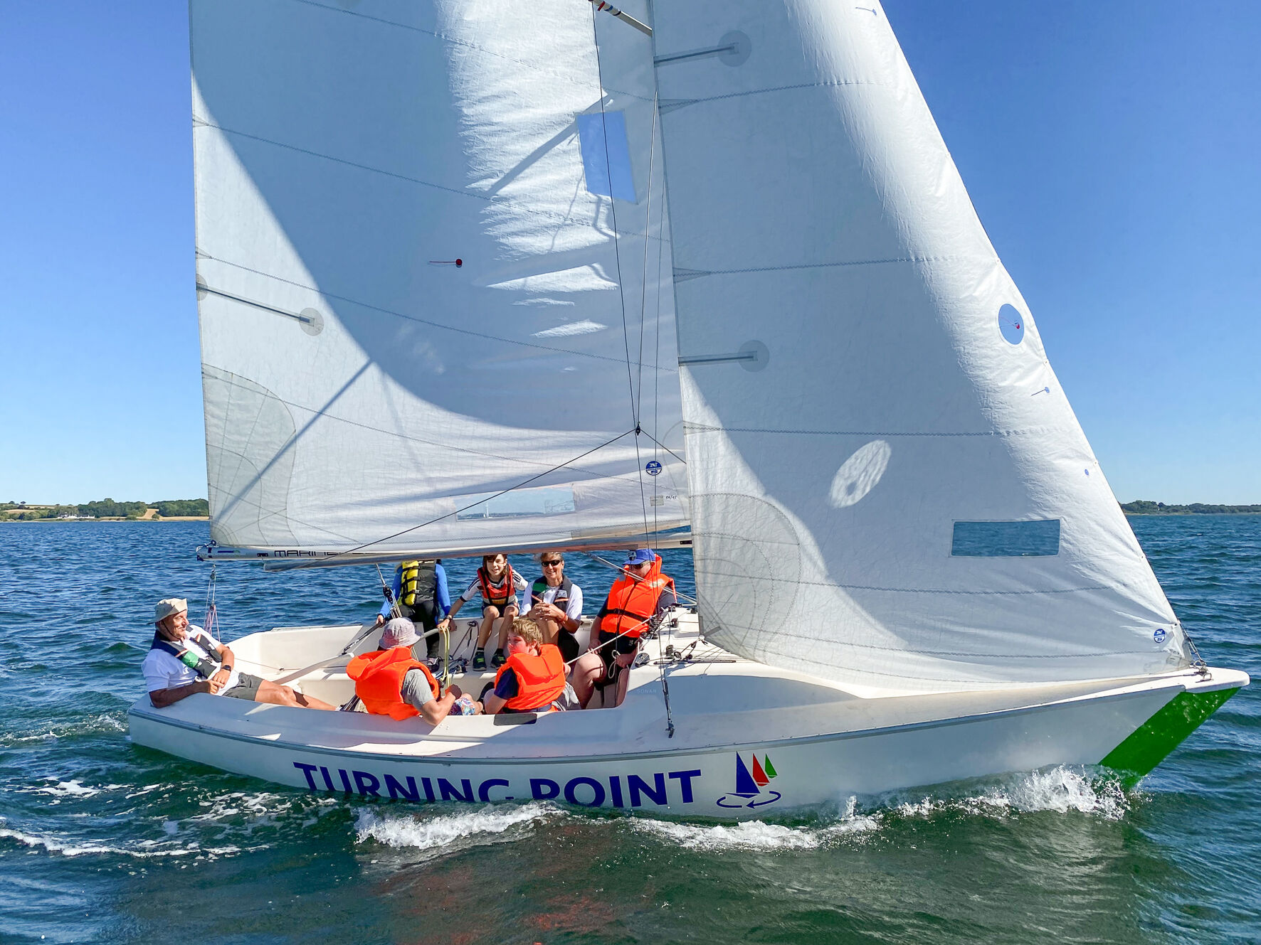 Eine Sonar segelt. Darin sitzen Jugendliche und ein Trainer. Alle tragen Rettungswesten.