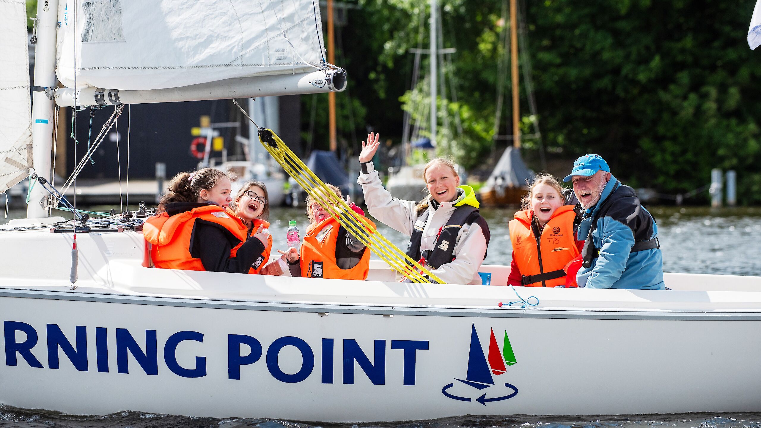 Children and coaches are sitting in a sailing boat. Two of them are waving.