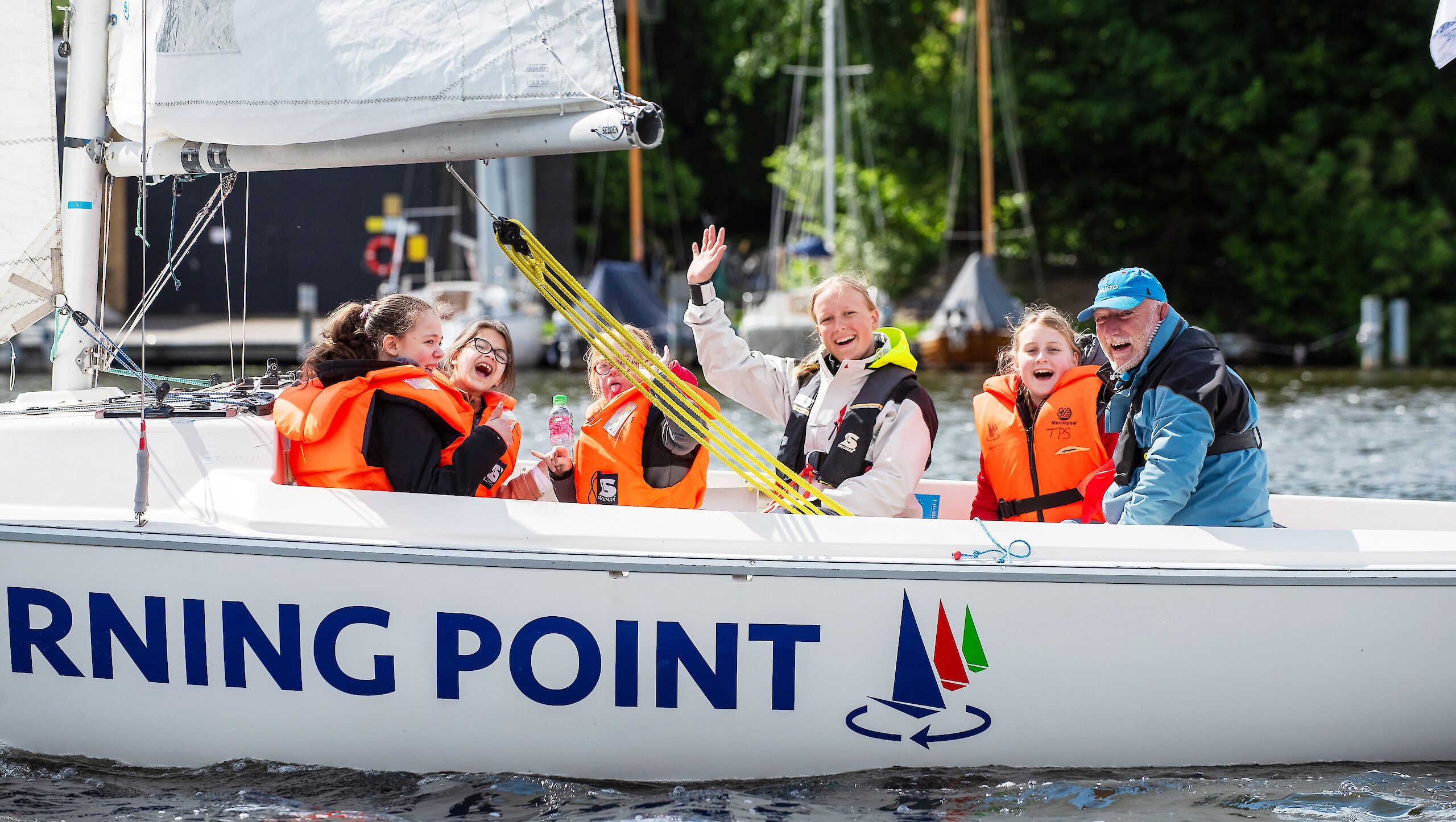 Vier Kinder und zwei Segeltrainierende sitzen auf einem Segelboot. Alle haben Schwimmwesten an und lachen in die Kamera.
