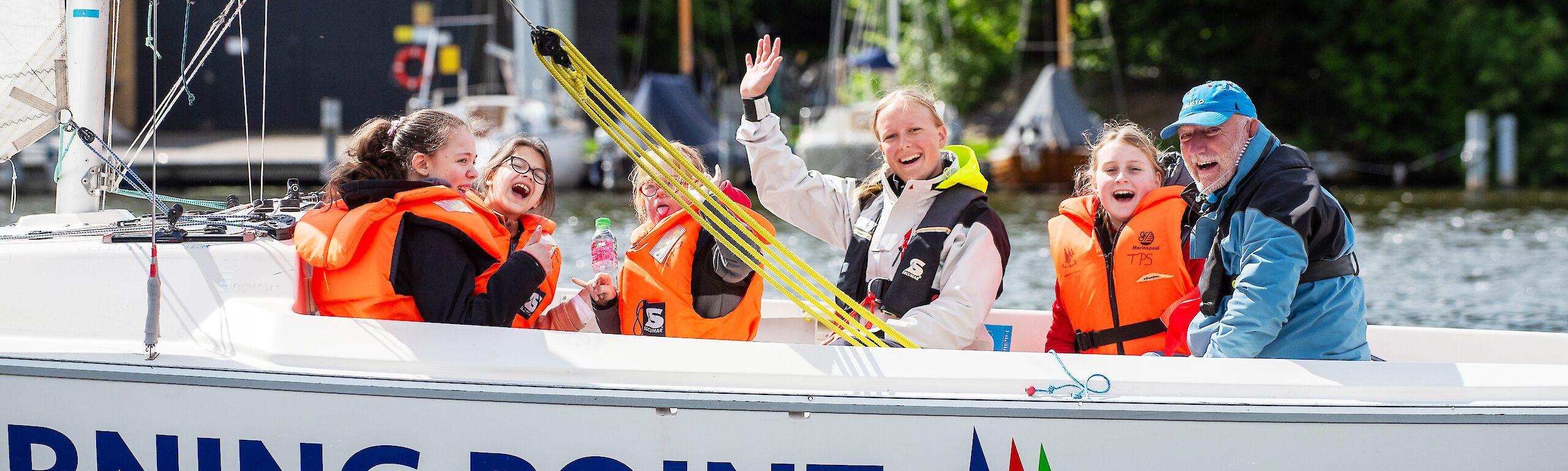 Vier Kinder und zwei Segeltrainierende sitzen auf einem Segelboot. Alle haben Schwimmwesten an und lachen in die Kamera.