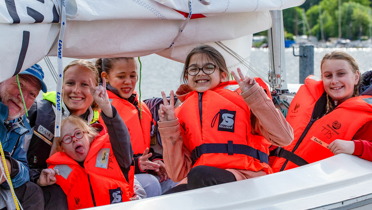 Vier Kinder und zwei Segeltrainierende sitzen auf einem Segelboot. Alle haben Schwimmwesten an und lachen in die Kamera.