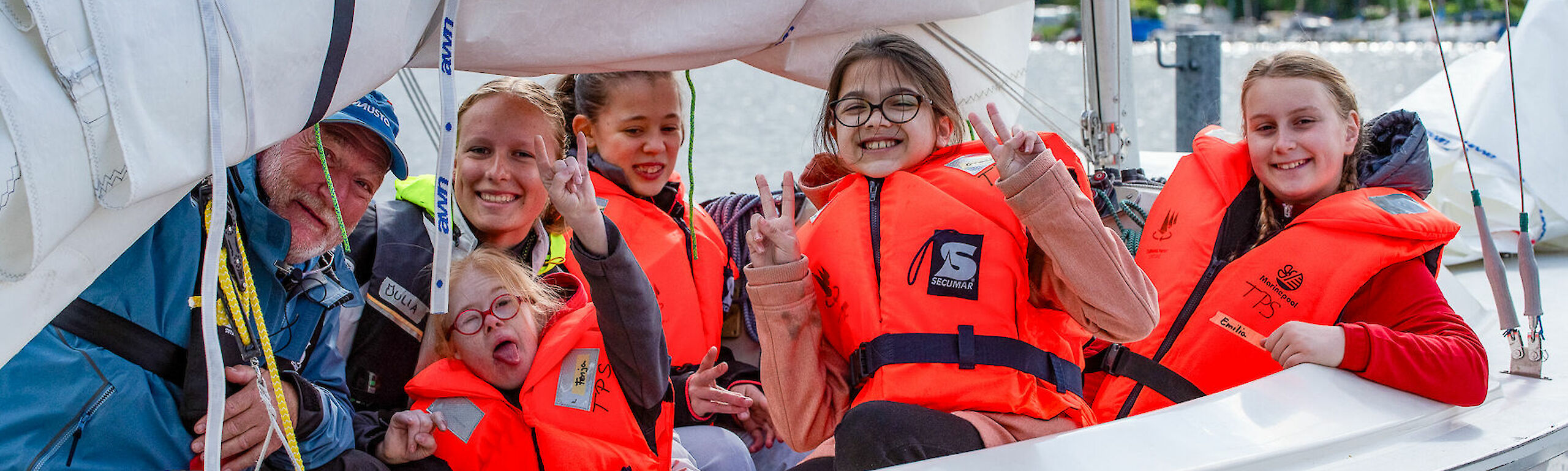 Vier Kinder und zwei Segeltrainierende sitzen auf einem Segelboot. Alle haben Schwimmwesten an und lachen in die Kamera.