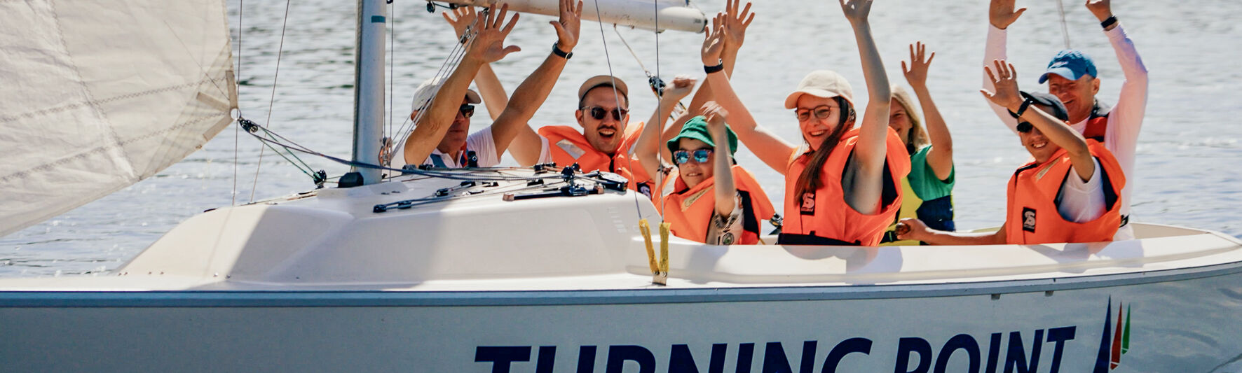 Menschen sitzen in einem Segelboot mit der Aufschrift Turning Point. Sie winken.