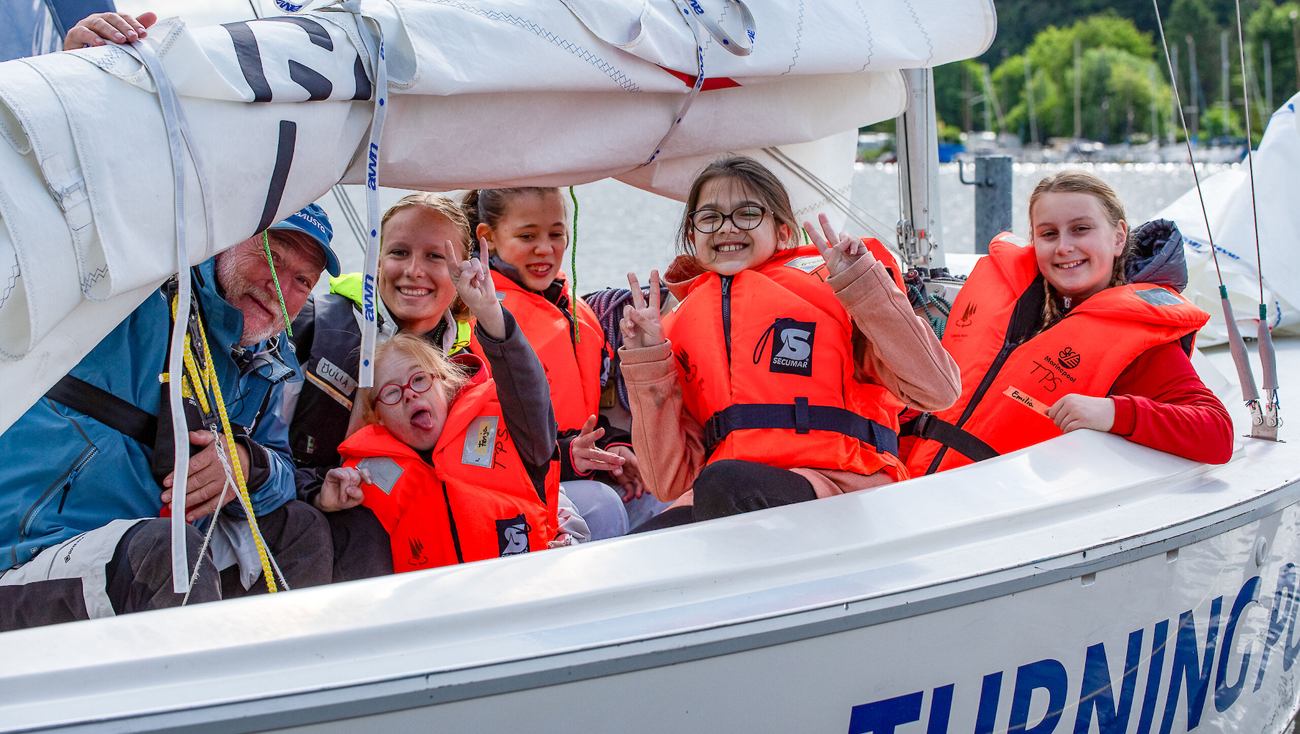 Vier Kinder und zwei Trainierende sitzen in einem Segelboot. Alle tragen Schwimmwesten und lachen.