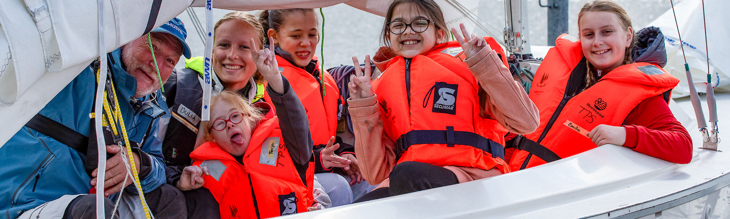 Vier Kinder und zwei Trainierende sitzen in einem Segelboot. Alle tragen Schwimmwesten und lachen.