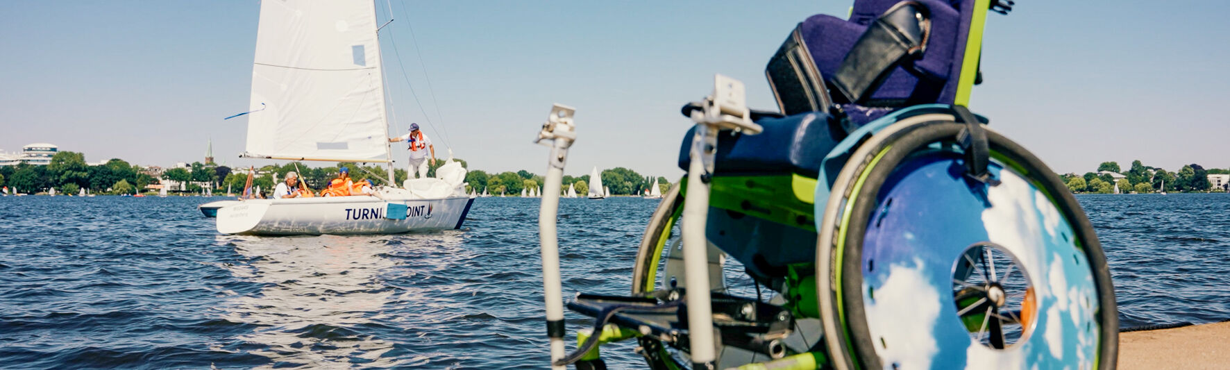 Ein Rollstuhl steht auf einem Steg. Im Hintergrund segelt ein Boot.