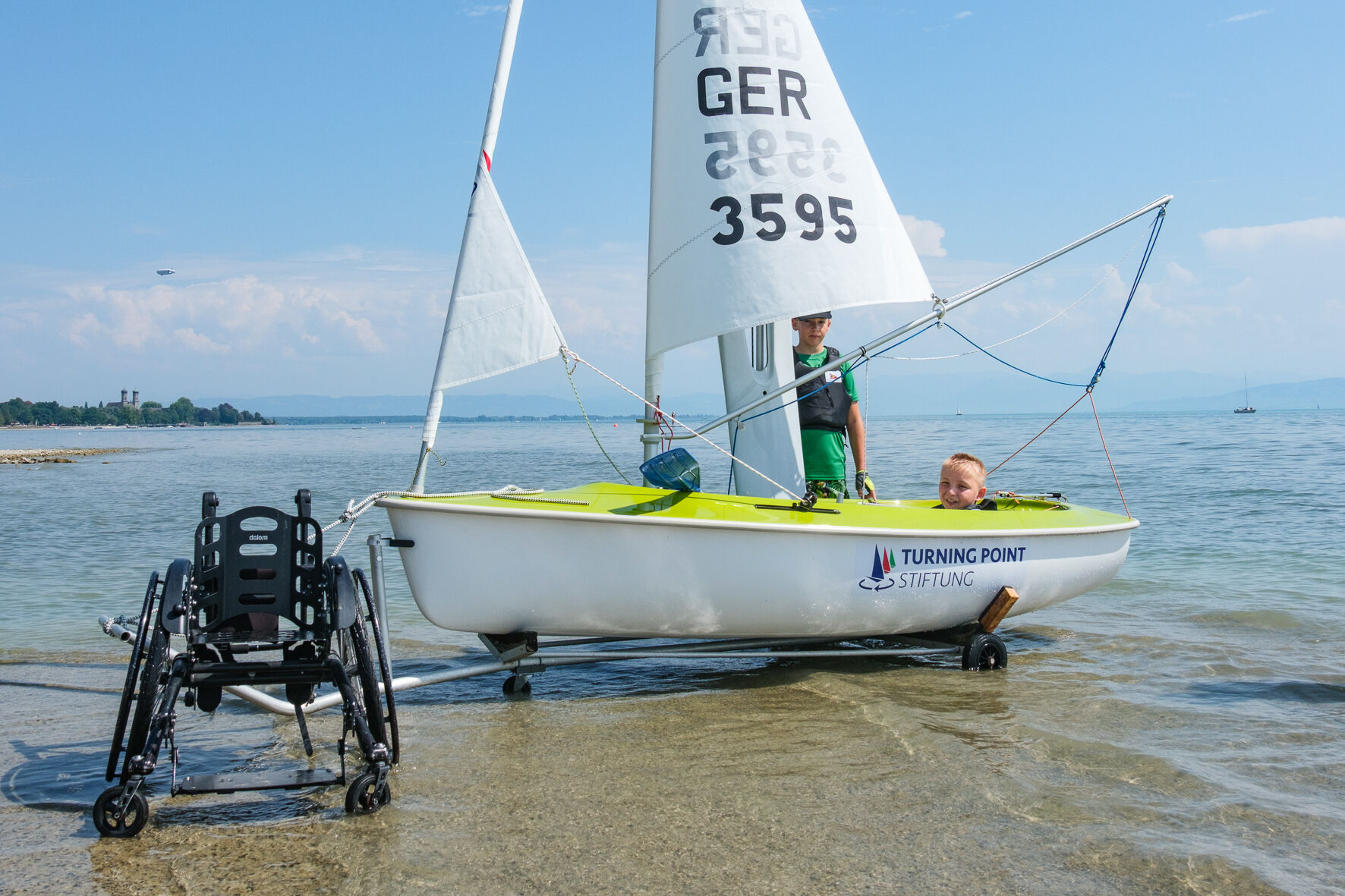Ein Rollstuhl steht neben einer Hansa 303. In dem Boot sitzt ein Kind.