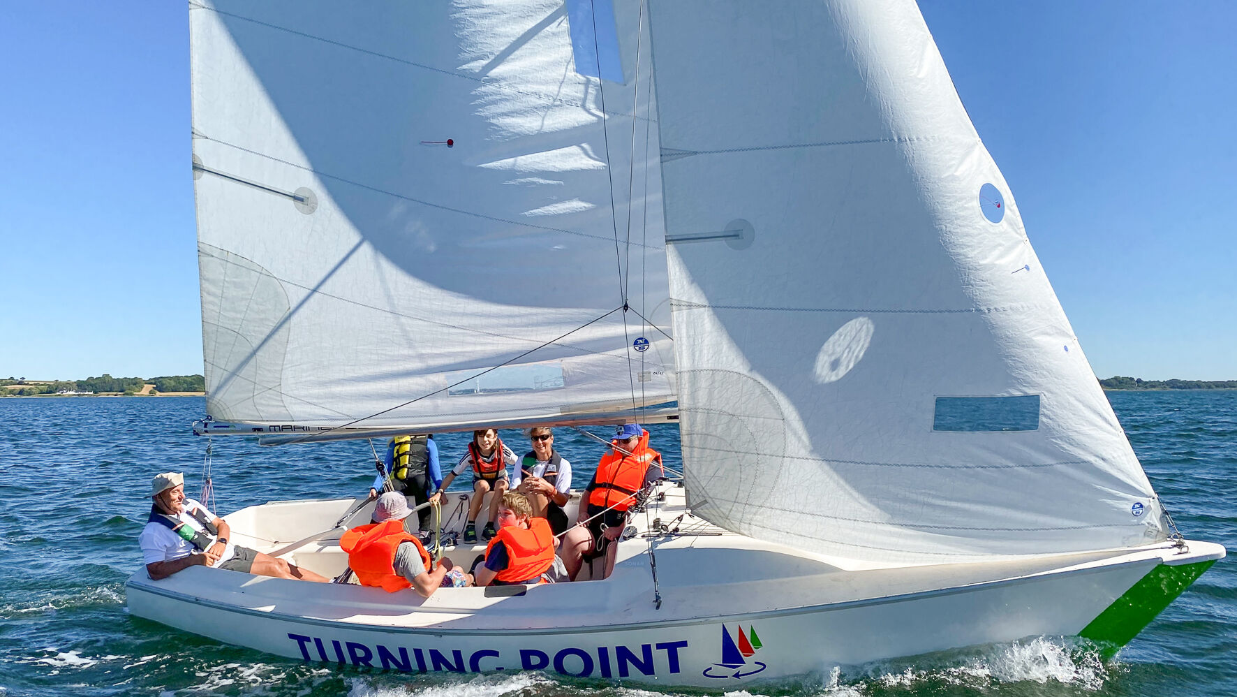 Menschen sitzen in einem Segelboot. Alle tragen Rettungswesten. Die Sonne scheint und das Boot segelt.