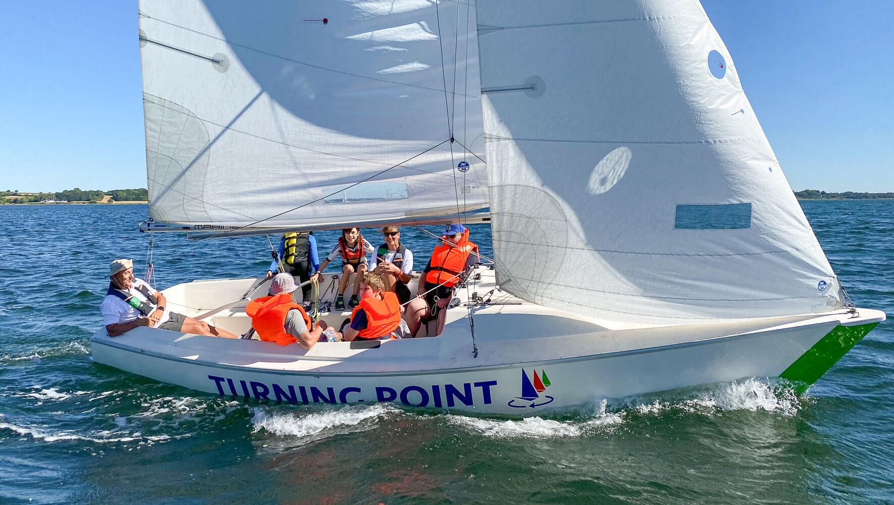 Menschen sitzen in einem Segelboot. Alle tragen Rettungswesten. Die Sonne scheint und das Boot segelt.
