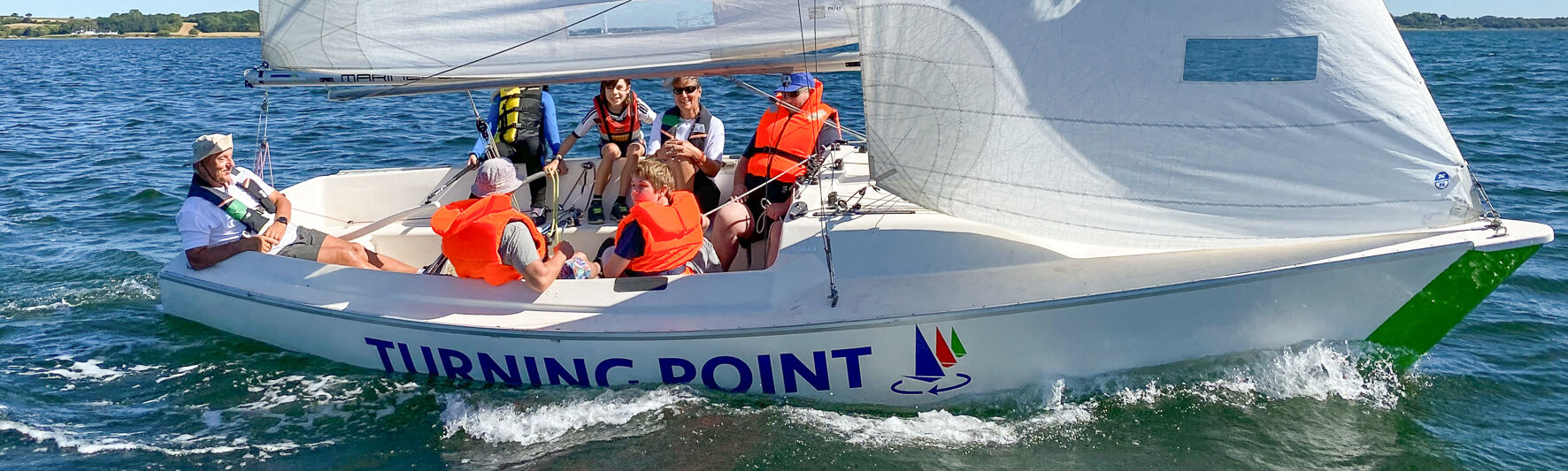 Menschen sitzen in einem Segelboot. Alle tragen Rettungswesten. Die Sonne scheint und das Boot segelt.