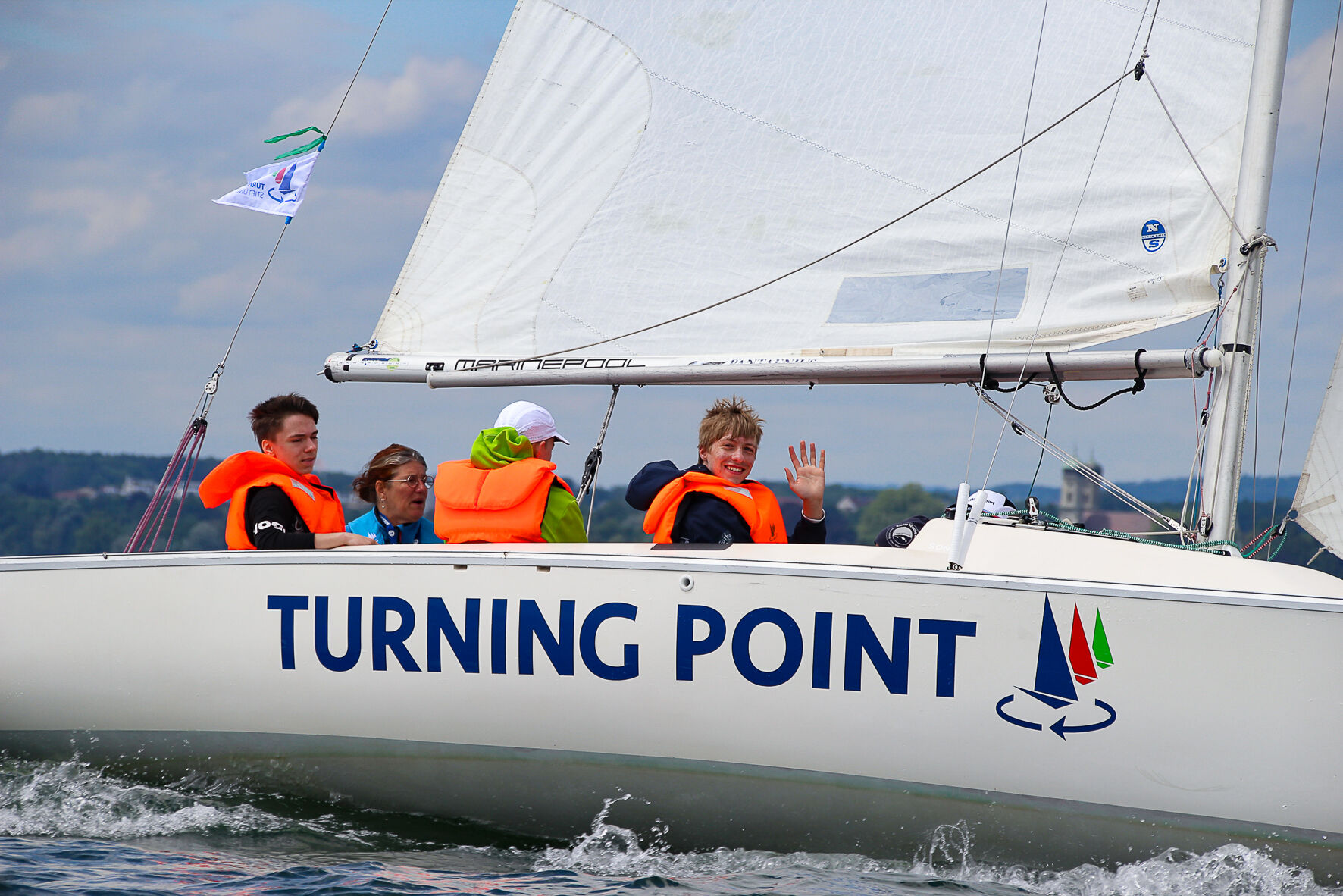 Jugendliche segeln auf einer Sonar. Einer lacht und winkt.