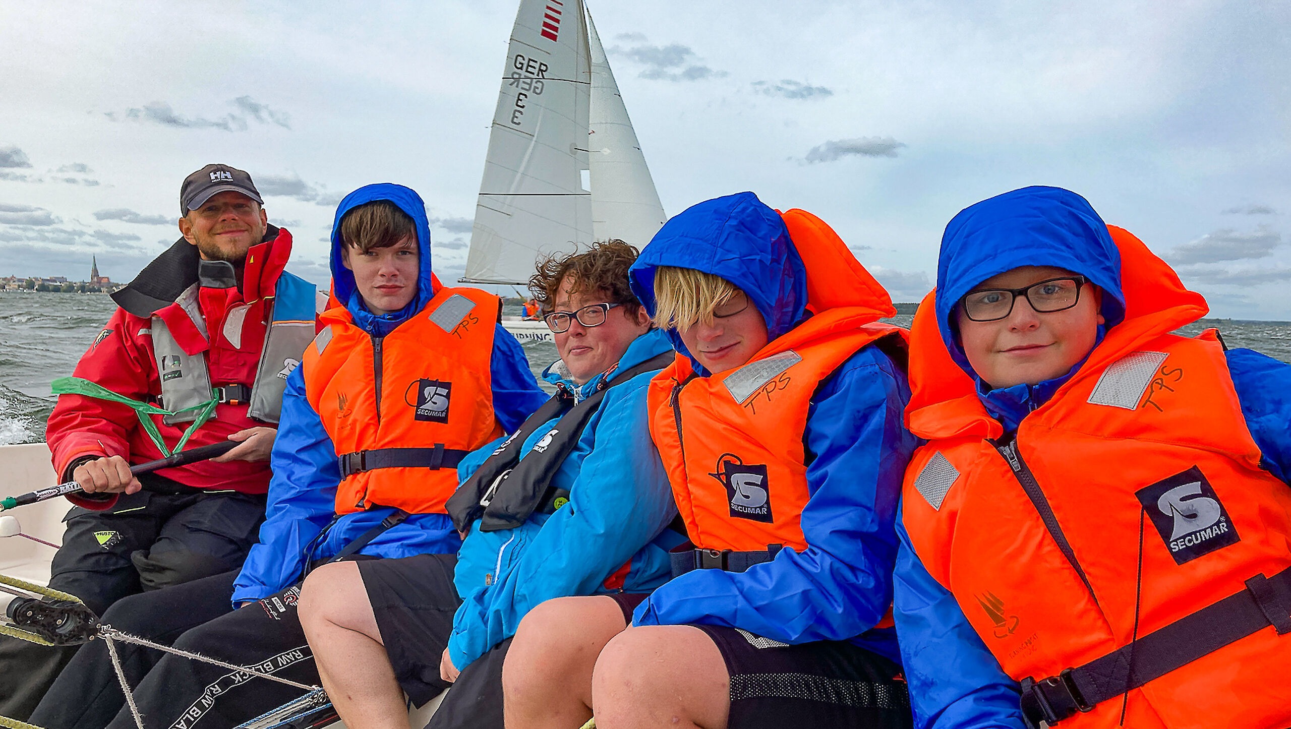 Fünf Personen sitzen in einem Segelboot, alle haben Schwimmwesten an