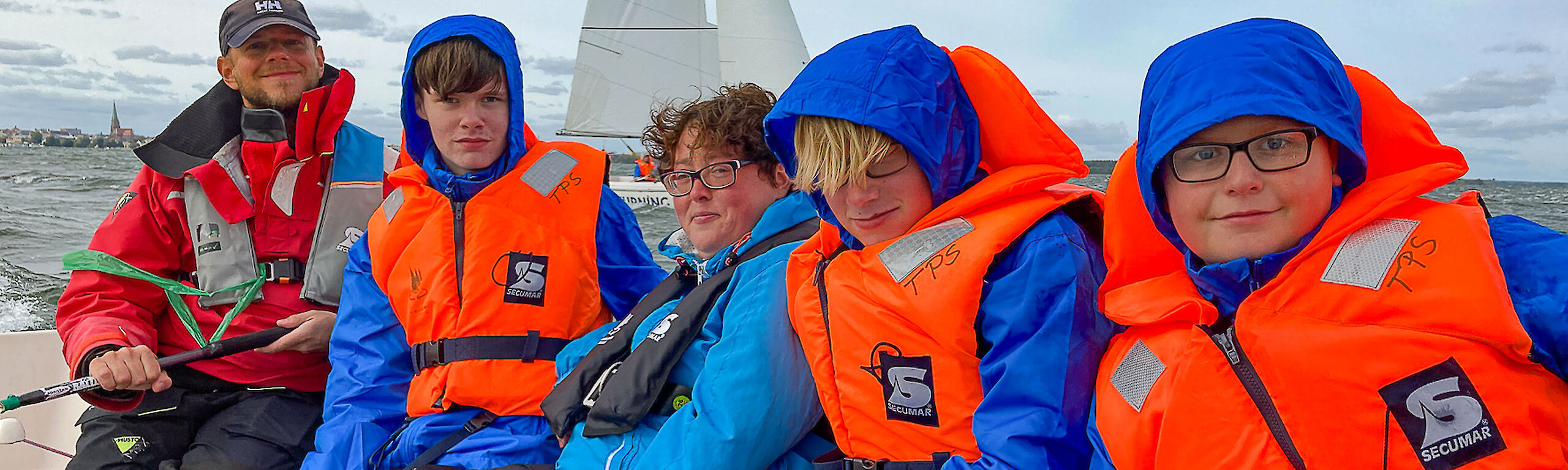 Fünf Personen sitzen in einem Segelboot, alle haben Schwimmwesten an
