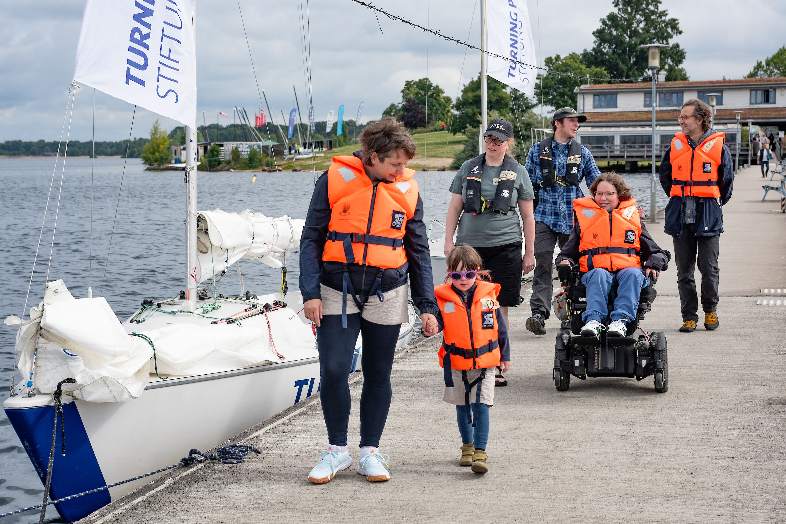 Menschen in Rettungswesten gehen über einen Bootssteg. Eine Person fährt mit einem Rollstuhl.